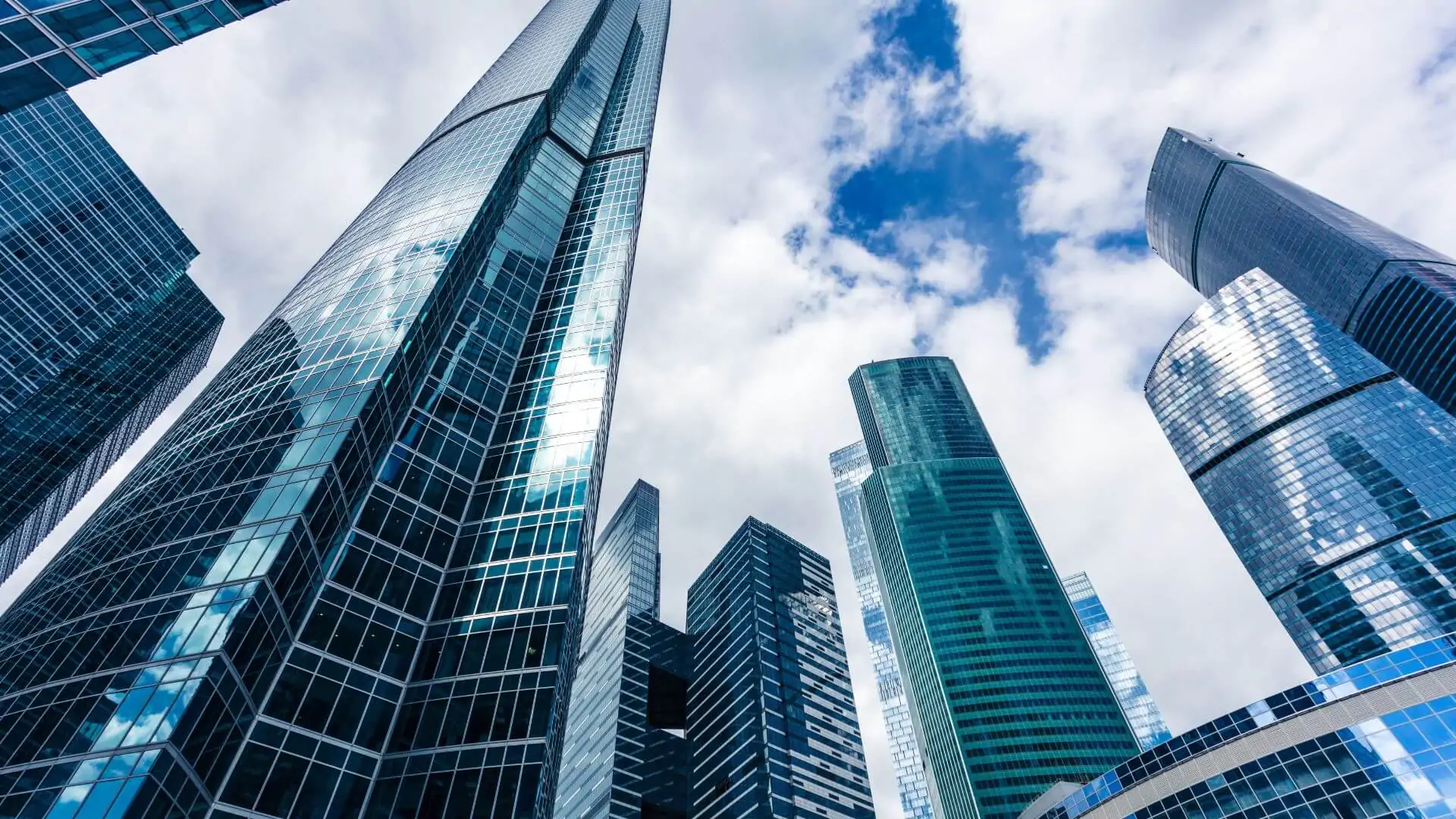 Vista de baixo para cima de modernos e imponentes arranha-céus corporativos espelhados contra um céu azul com nuvens. Simboliza a solidez e o alto nível do mercado de capitais.